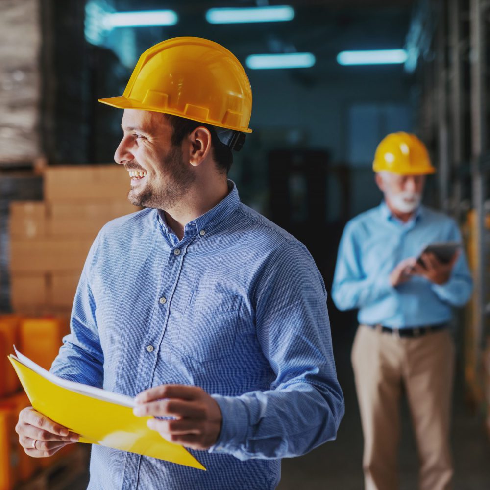 Young smiling Caucasian businessman holding folder with documents and looking away. In background older businessman using tablet. Warehouse interior.